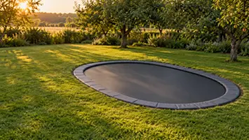Stor oval mørkegrå trampolin gravet ned i en åben græsplæne med æbletræer i god afstand i gyldent aftenlys